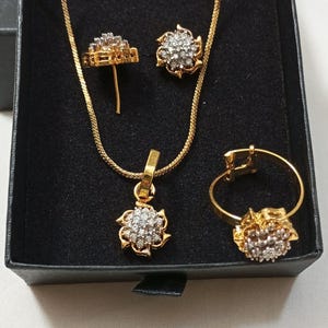 May include: A gold-toned jewelry set consisting of a necklace with a floral-shaped pendant, a pair of matching stud earrings, and a ring. Each piece features a central cluster of small, clear, sparkling stones set within a gold-colored metal frame.