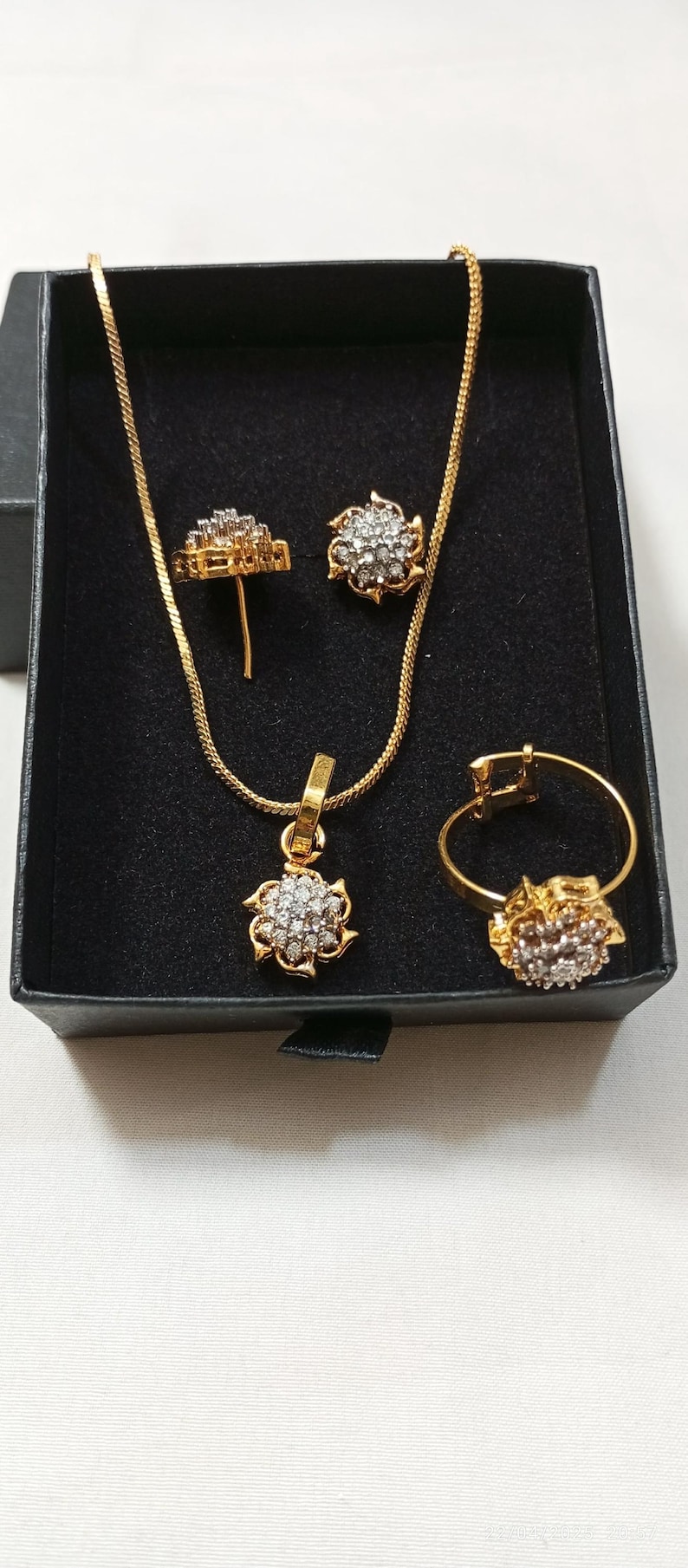 May include: A gold-toned jewelry set consisting of a necklace with a floral-shaped pendant, a pair of matching stud earrings, and a ring. Each piece features a central cluster of small, clear, sparkling stones set within a gold-colored metal frame.