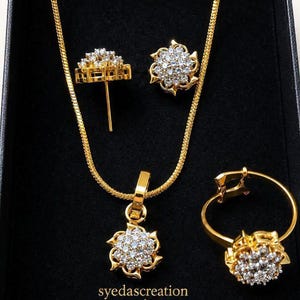 May include: A gold-toned jewelry set featuring a necklace with a floral pendant, a pair of stud earrings, and a ring, all adorned with clear stones. The pieces are arranged in a black box with the text syedascreation printed in gold lettering.