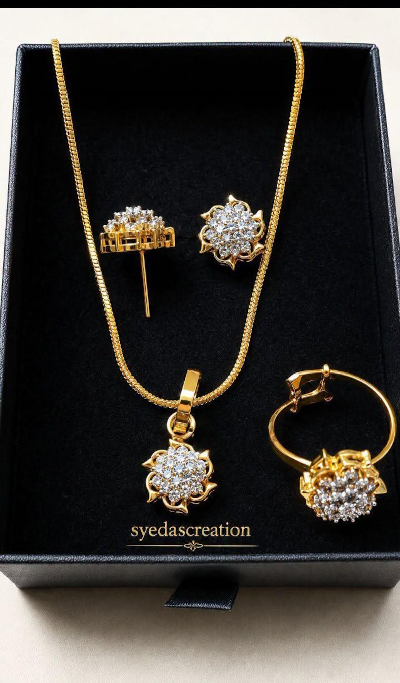 May include: A gold-toned jewelry set featuring a necklace with a floral pendant, a pair of stud earrings, and a ring, all adorned with clear stones. The pieces are arranged in a black box with the text syedascreation printed in gold lettering.