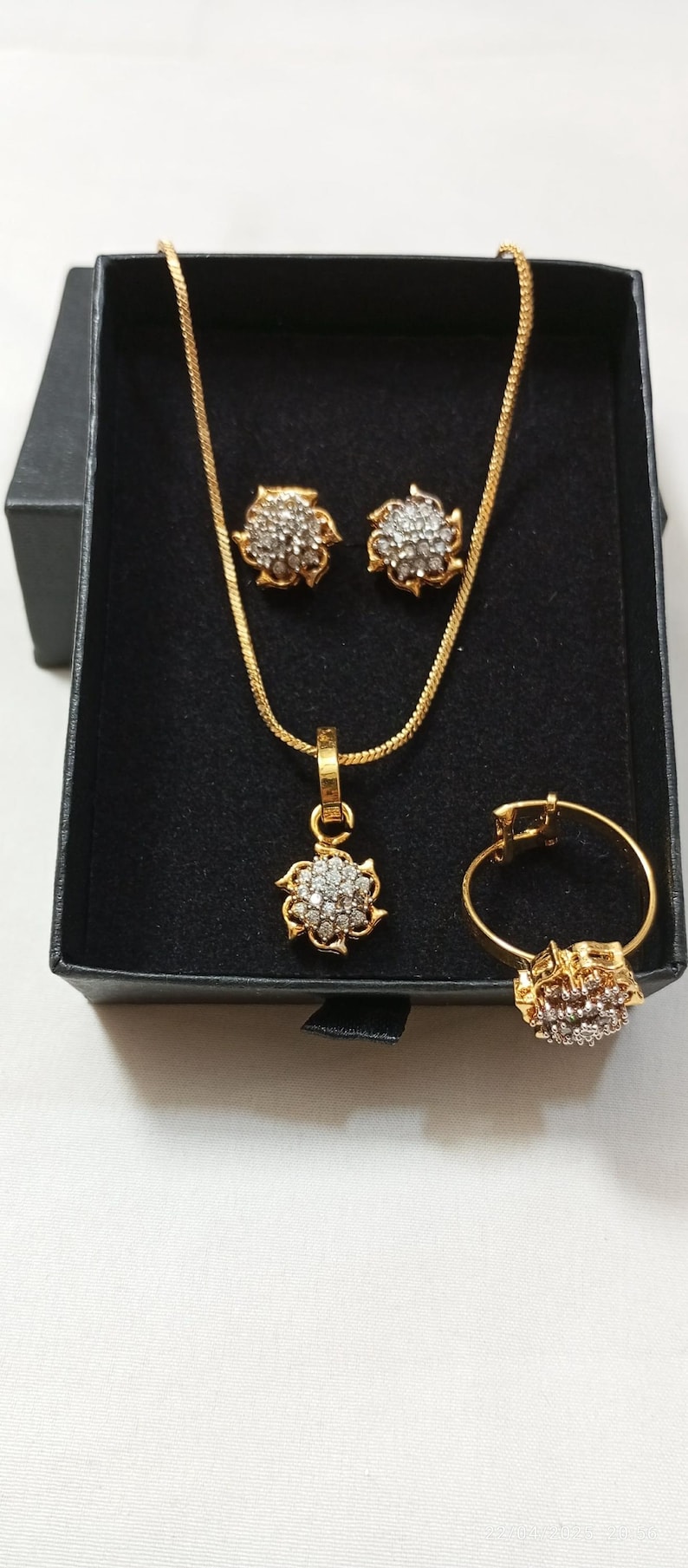 May include: A gold-toned jewelry set consisting of a necklace with a floral-shaped pendant, a pair of matching stud earrings, and a ring. Each piece features a cluster of small, clear, sparkling stones set within a gold-colored metal frame.