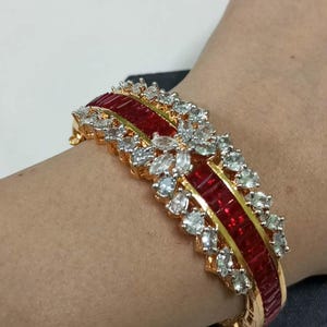 Peut inclure: Bracelet doré avec une bande centrale rouge et des pierres claires et scintillantes. Le bracelet présente un motif répétitif de pierres rouges rectangulaires et de pierres claires taillées en rond. Un bijou luxueux.