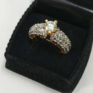 May include: A gold ring with a large, clear center stone and smaller stones set in a double band. The ring is displayed in a black velvet ring box.