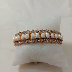 May include: A gold-toned bracelet featuring a row of oval, white gemstones, interspersed with smaller, clear stones. The bracelet is displayed on a white, textured surface, showcasing its intricate design and elegant appeal. This piece of jewelry is suitable for special occasions.