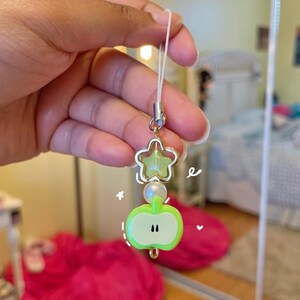 May include: A charm with a green apple and star design. The apple charm has a light green color with black accents. The star charm is gold with a light green center. A white string is attached to the charm.