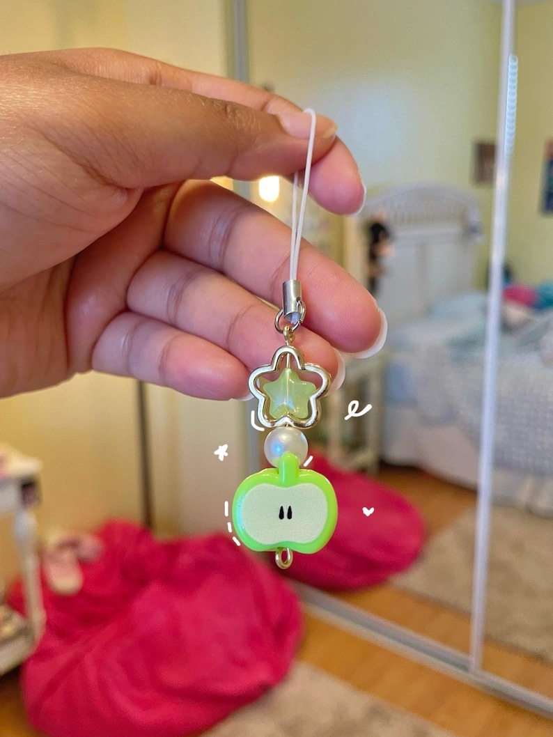 May include: A charm with a green apple and star design. The apple charm has a light green color with black accents. The star charm is gold with a light green center. A white string is attached to the charm.