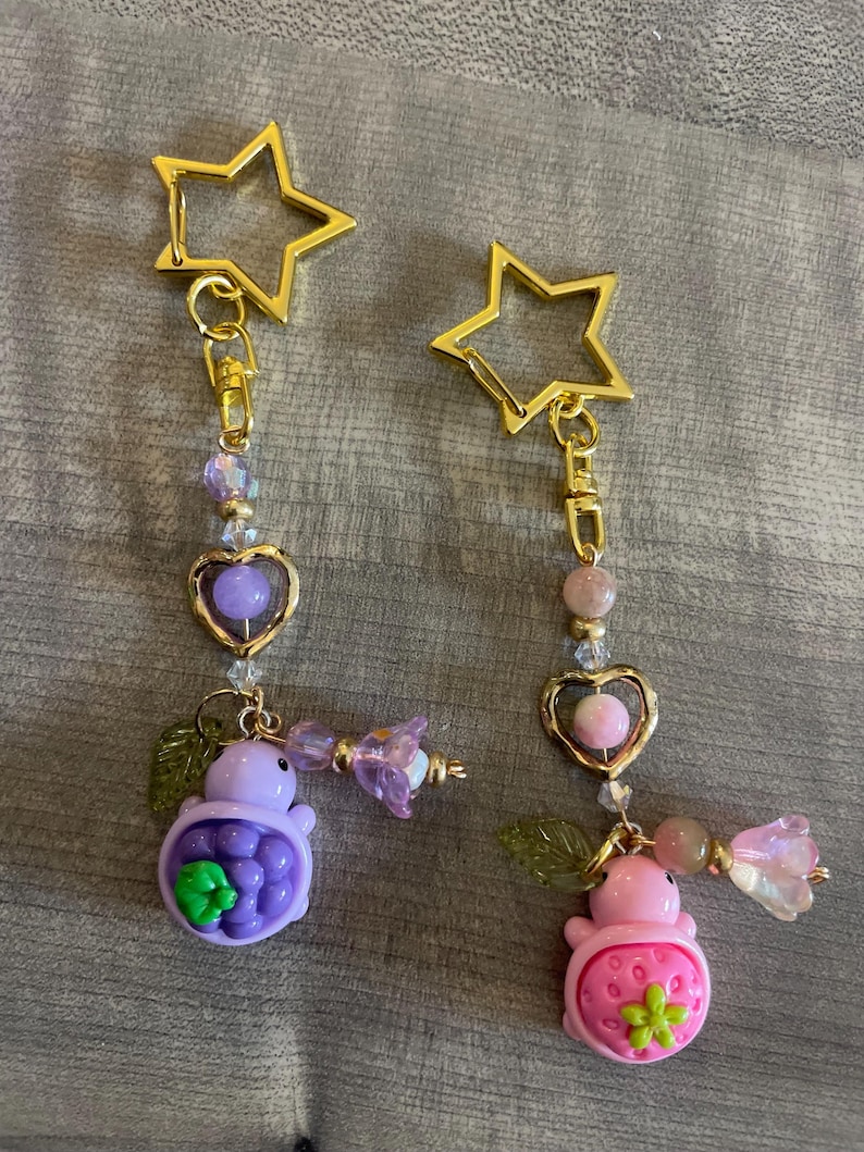 May include: Two keychains with gold star clasps. One keychain features a purple turtle with a green flower, the other a pink strawberry turtle with a green flower. Both have heart-shaped charms and beaded accents.