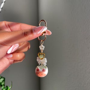 May include: A gold-colored keychain with a lobster clasp, adorned with a white flower, a gold heart, a white bow, and a ceramic frog charm. The frog charm is white with orange and green accents.