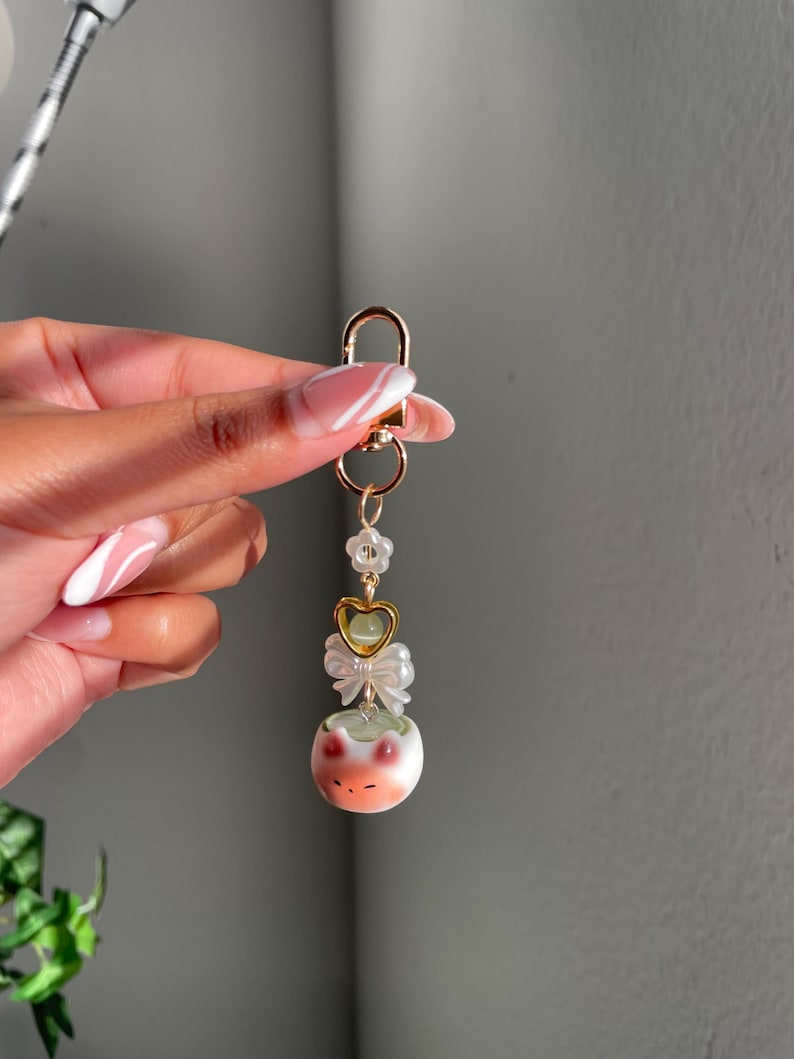 May include: A gold-colored keychain with a lobster clasp, adorned with a white flower, a gold heart, a white bow, and a ceramic frog charm. The frog charm is white with orange and green accents.