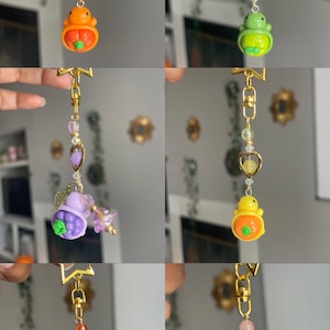 May include: A collection of six keychains with gold-colored hardware. Each keychain features a charm with a unique design, including a pumpkin, a frog, a berry, a strawberry, and a pink strawberry. The keychains are adorned with beads and heart-shaped accents.