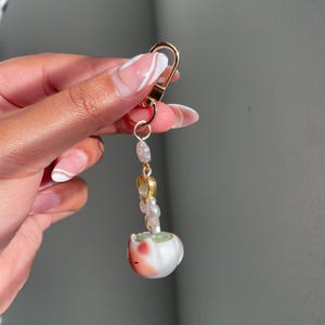 May include: A gold-colored keychain with a lobster clasp. The keychain features a string of small, iridescent beads and a small, white ceramic cat figurine with red and pink accents.
