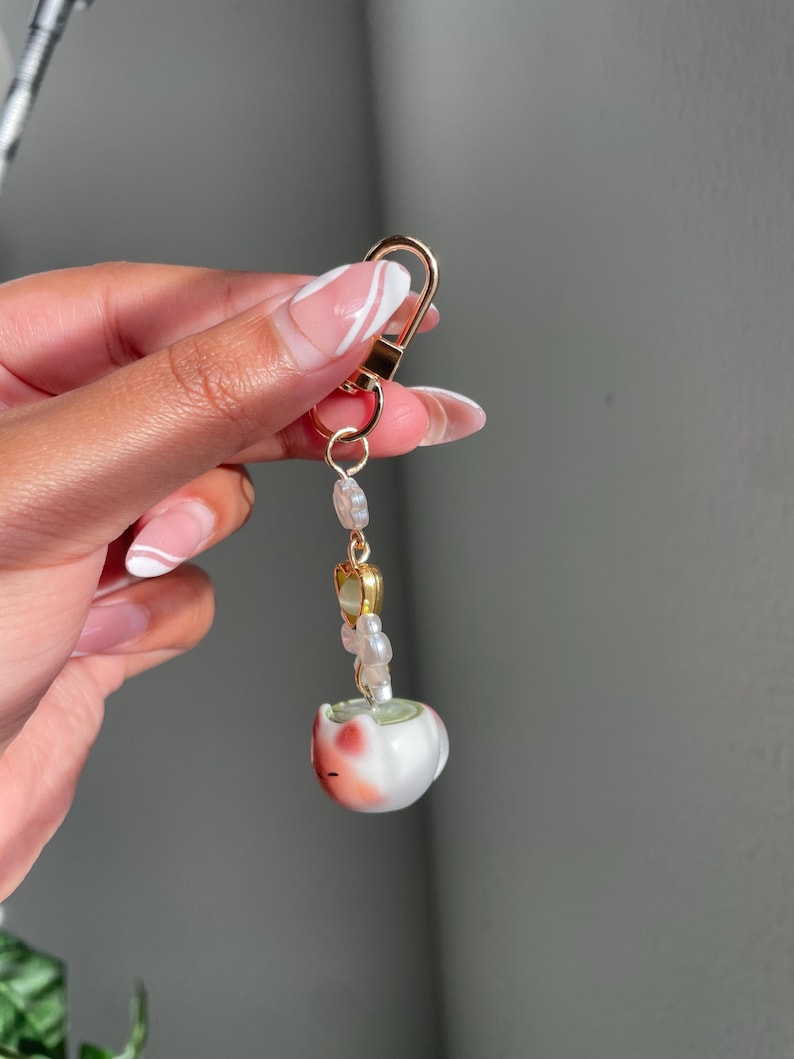 May include: A gold-colored keychain with a lobster clasp. The keychain features a string of small, iridescent beads and a small, white ceramic cat figurine with red and pink accents.