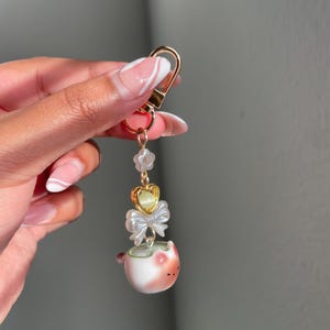 May include: A gold-colored keychain with a decorative charm. The charm features a small, white and pink ceramic cup with a bow, a heart-shaped accent, and a string of clear beads. The keychain is held by a hand with manicured nails.