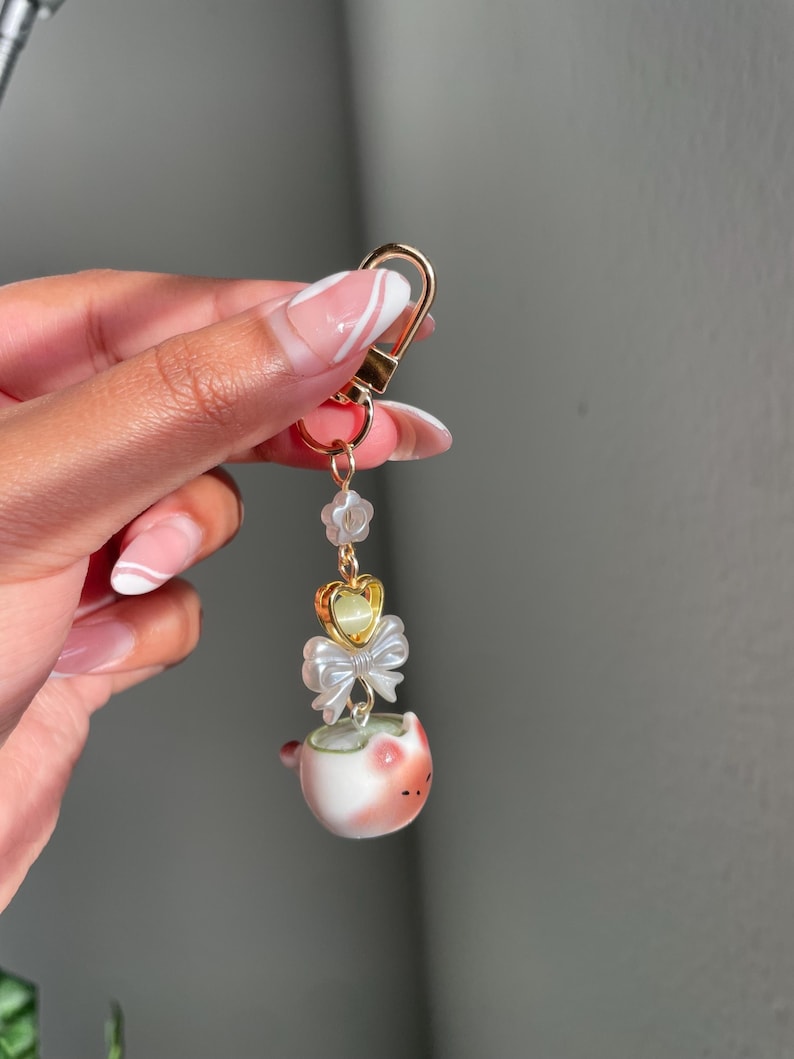 May include: A gold-colored keychain with a decorative charm. The charm features a small, white and pink ceramic cup with a bow, a heart-shaped accent, and a string of clear beads. The keychain is held by a hand with manicured nails.