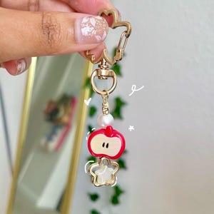 May include: A gold-colored keychain with a star-shaped clasp and a charm featuring a red apple and a star. The apple charm has a white interior and a pearl bead. The keychain is held up by a hand.