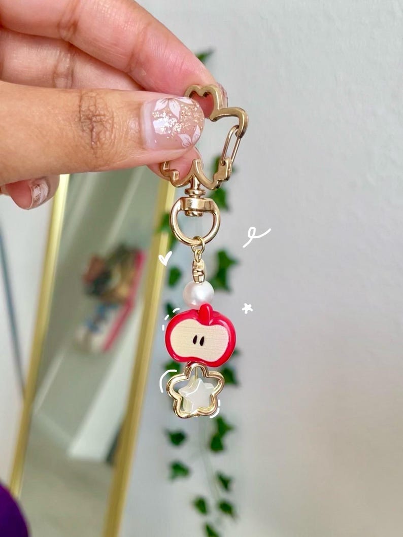May include: A gold-colored keychain with a star-shaped clasp and a charm featuring a red apple and a star. The apple charm has a white interior and a pearl bead. The keychain is held up by a hand.