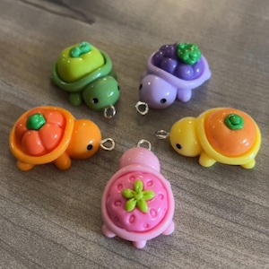 May include: Five colorful turtle charms with fruit-themed shells. The turtles are in shades of green, purple, orange, yellow, and pink. Each charm has a small metal loop for attaching to jewelry or other crafts.