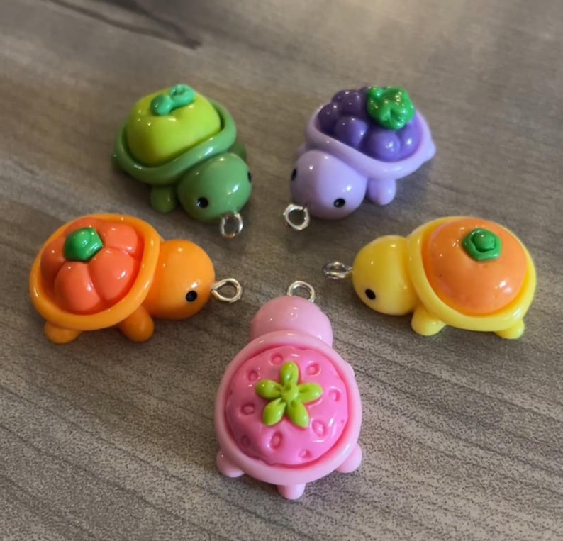 May include: Five colorful turtle charms with fruit-themed shells. The turtles are in shades of green, purple, orange, yellow, and pink. Each charm has a small metal loop for attaching to jewelry or other crafts.