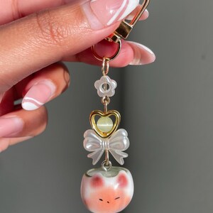 May include: A gold-colored keychain with a lobster clasp, featuring a flower, heart, bow, and a small, round, white and orange cat-like charm. The charm has a painted face with closed eyes and red cheeks.