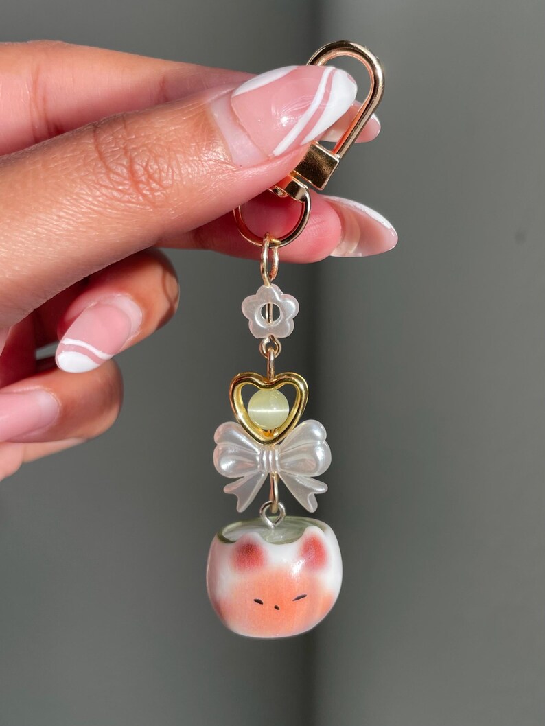 May include: A gold-colored keychain with a lobster clasp, featuring a flower, heart, bow, and a small, round, white and orange cat-like charm. The charm has a painted face with closed eyes and red cheeks.