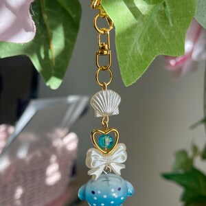 May include: A gold-coloured keychain featuring a seashell, a heart-shaped charm with a blue bead, a white bow, and a blue whale-like figure with white spots. The keychain is suspended against a backdrop of green leaves and pink flowers.