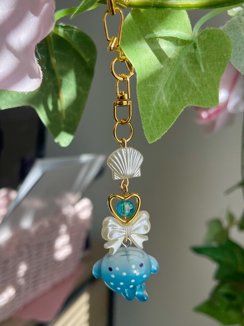 May include: A gold-coloured keychain featuring a seashell, a heart-shaped charm with a blue bead, a white bow, and a blue whale-like figure with white spots. The keychain is suspended against a backdrop of green leaves and pink flowers.