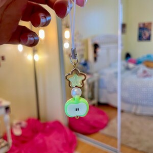 May include: A charm with a green apple, a star, and a pearl bead. The apple charm has a white outline and two black dots. The star is a light green color. The charm is attached to a white string.