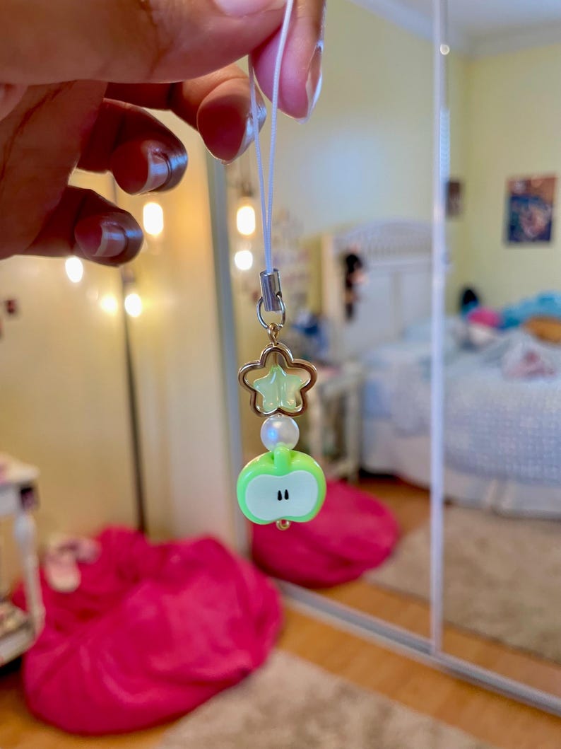 May include: A charm with a green apple, a star, and a pearl bead. The apple charm has a white outline and two black dots. The star is a light green color. The charm is attached to a white string.