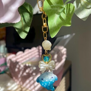 May include: A gold-coloured chain with a charm featuring a blue and white spotted dolphin, a white bow, a heart-shaped charm with a blue bead, and a seashell. The charm is hanging from a gold-coloured clasp.