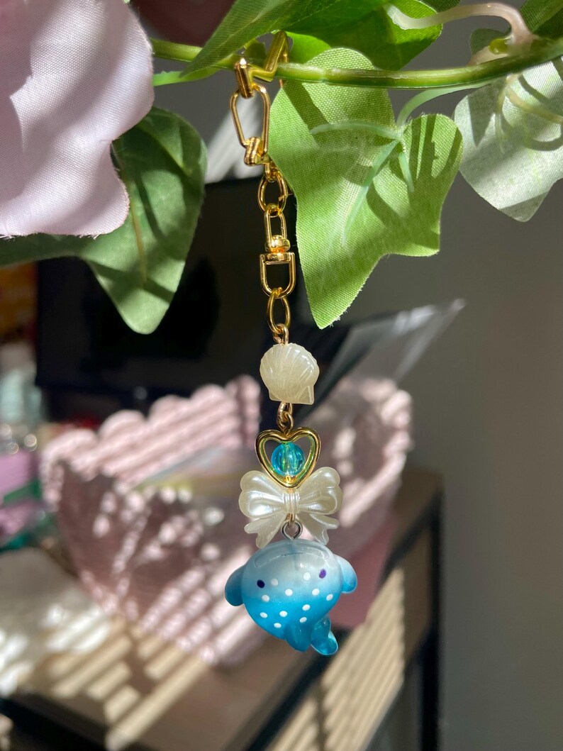 May include: A gold-coloured chain with a charm featuring a blue and white spotted dolphin, a white bow, a heart-shaped charm with a blue bead, and a seashell. The charm is hanging from a gold-coloured clasp.