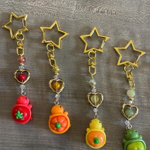 May include: Four keychains with gold star-shaped clasps. Each keychain has a different colored turtle charm: red, orange, yellow, and green. The charms are attached to gold chains with heart-shaped and beaded accents. The keychains are on a gray surface.