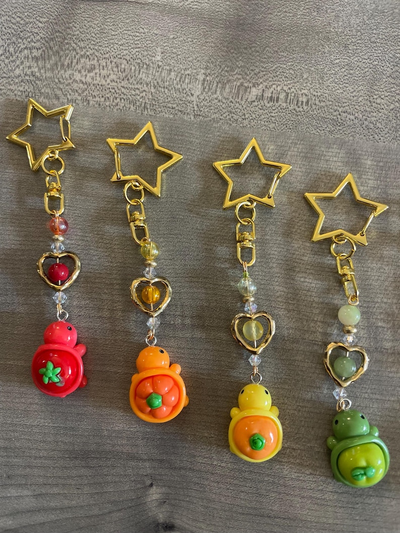 May include: Four keychains with gold star-shaped clasps. Each keychain has a different colored turtle charm: red, orange, yellow, and green. The charms are attached to gold chains with heart-shaped and beaded accents. The keychains are on a gray surface.