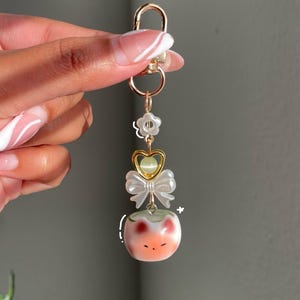 May include: A gold-colored keychain with a lobster clasp. The keychain features a flower, a heart-shaped charm, a white bow, and a peach-colored cat-shaped charm with red accents. The keychain is held by a person.