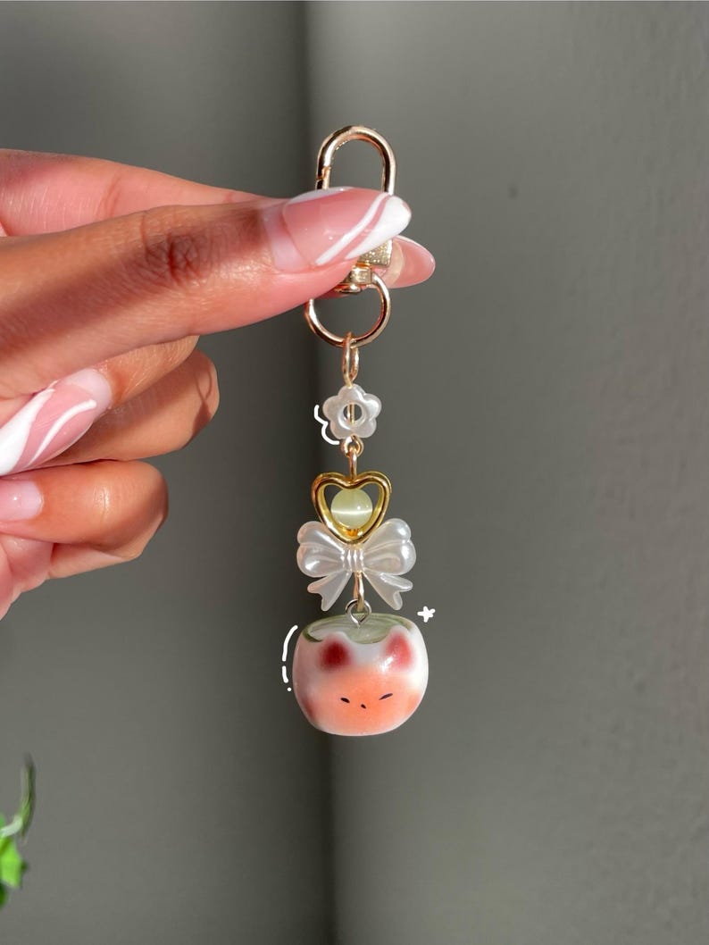 May include: A gold-colored keychain with a lobster clasp. The keychain features a flower, a heart-shaped charm, a white bow, and a peach-colored cat-shaped charm with red accents. The keychain is held by a person.