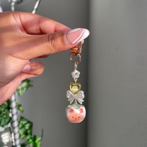 May include: A gold-colored keychain with a decorative charm. The charm features a small, round, peach-colored object with a cat face design, a white bow, and a gold heart. The keychain is attached to a gold clasp.