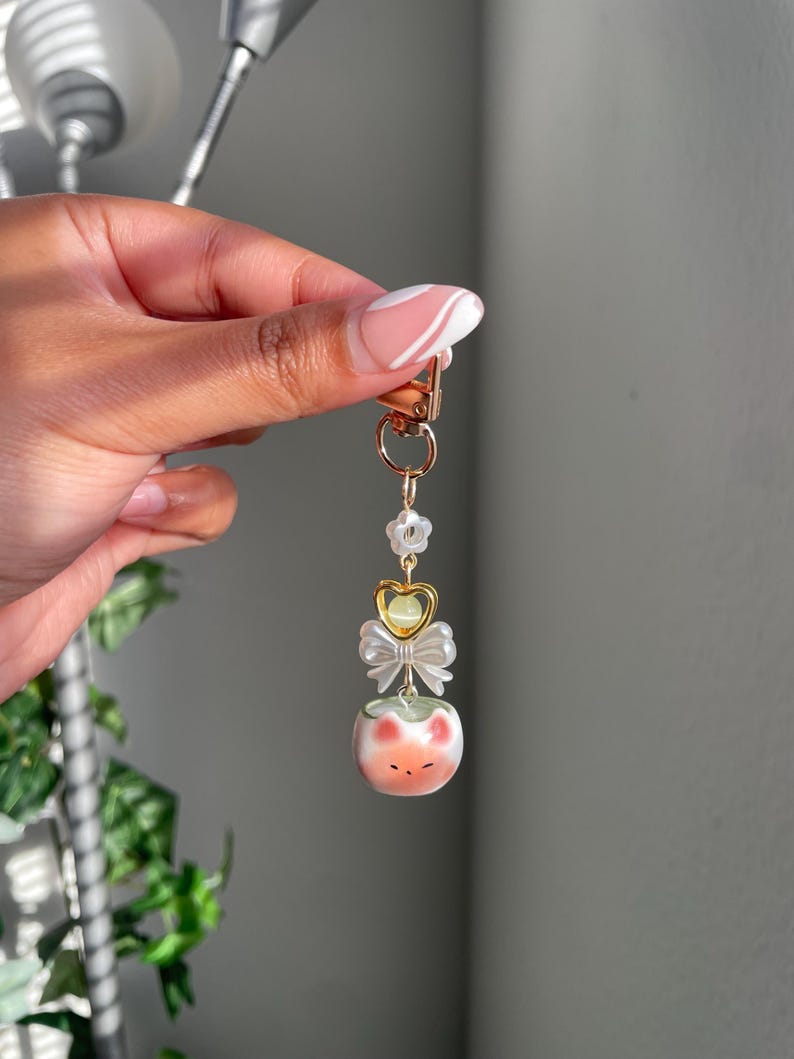 May include: A gold-colored keychain with a decorative charm. The charm features a small, round, peach-colored object with a cat face design, a white bow, and a gold heart. The keychain is attached to a gold clasp.