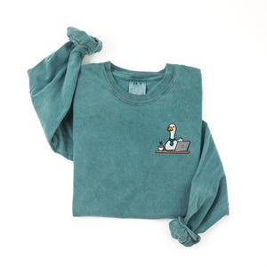 May include: A teal long-sleeve crewneck sweatshirt featuring an embroidered graphic on the left chest. The graphic depicts a white goose wearing a blue necktie, sitting at a desk with a laptop and a steaming cup of coffee on a brown surface.