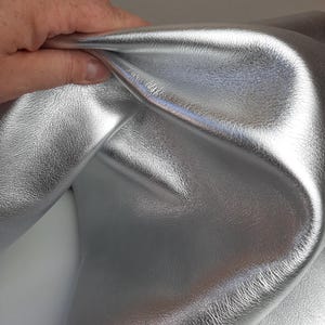 May include: Close-up of a piece of silver metallic leather. The material is folded, revealing its shiny, textured surface. The leather has a smooth, reflective quality, suggesting it could be used for fashion or craft projects.