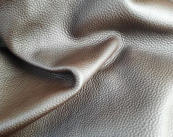 May include: Close-up of a piece of metallic silver leather with a textured surface. The leather is folded, creating shadows and highlights that emphasize its supple quality. The material has a subtle sheen, suggesting a high-quality finish.