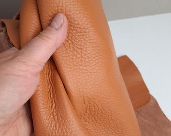 May include: Close-up of a piece of orange-brown leather with a textured surface. The leather is being held, showing its flexibility and the grain of the material. A smaller piece of leather is visible in the background.