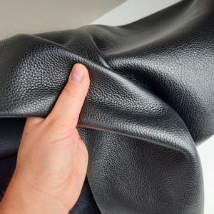 Peut inclure: Gros plan sur un morceau de cuir noir à surface texturée. Une main tient le cuir, soulignant sa souplesse et sa qualité. Le cuir semble doux et souple, adapté à diverses utilisations.