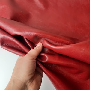 Op de afbeelding: Een close-up van een stuk rood leer. Het leer is glad en heeft een lichte glans. De textuur van het leer is zichtbaar. Het leer wordt door een hand vastgehouden.