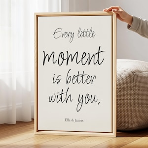 May include: A rectangular wall art piece in a light-colored wooden frame featuring a white background with black text that reads: Every little moment is better with you. Below the main text, the names Ella & James are printed in a smaller font.