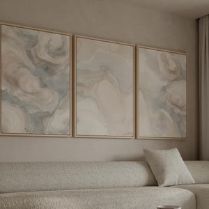 May include: Three framed abstract art pieces with swirling patterns in neutral tones of beige, gray, and white. The artwork is displayed above a light-colored sofa with a matching pillow, creating a cohesive and modern interior design.