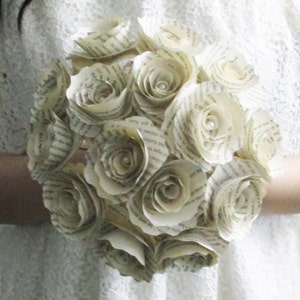 May include: A bouquet of paper roses crafted from book pages. The roses are white and have a delicate, vintage appearance. Each rose features a small pearl at its centre.