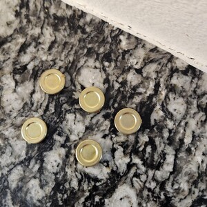 Può includere: Cinque piccoli coperchi rotondi in metallo dorato sono sparsi su una superficie screziata grigia e bianca. I coperchi hanno un design poco profondo a forma di piatto con un bordo leggermente rialzato. Lo sfondo include un tessuto bianco.
