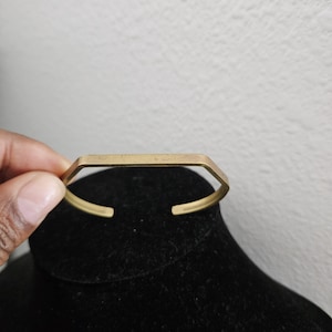 May include: A gold-colored cuff bracelet with a geometric design. The bracelet has a rectangular top and open ends. The metal has a slightly aged appearance. The bracelet is displayed on a black surface.
