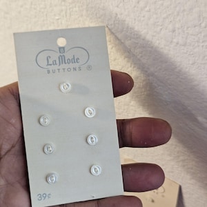 May include: A card of six clear, round La Mode Buttons with a textured design. The card is light gray with the brand name and the price 39¢ printed on it.