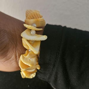 May include: A seashell bracelet featuring a spiral design. The bracelet is made of cream-colored seashells and small, round, white beads. The shells are arranged in a stacked pattern, creating a unique and natural look. A perfect accessory for beach lovers.
