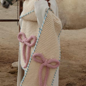May include: Cream-coloured knitted scarf with light blue trim and two pink bow accents. The scarf is rectangular and appears to be handmade. The background includes a horse and a fence.